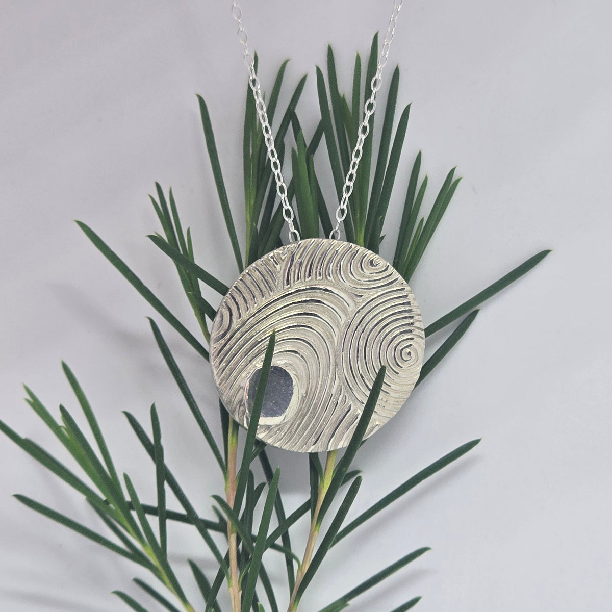 Fine silver tidal spiral pendant laid on white surface with rosemary sprigs
